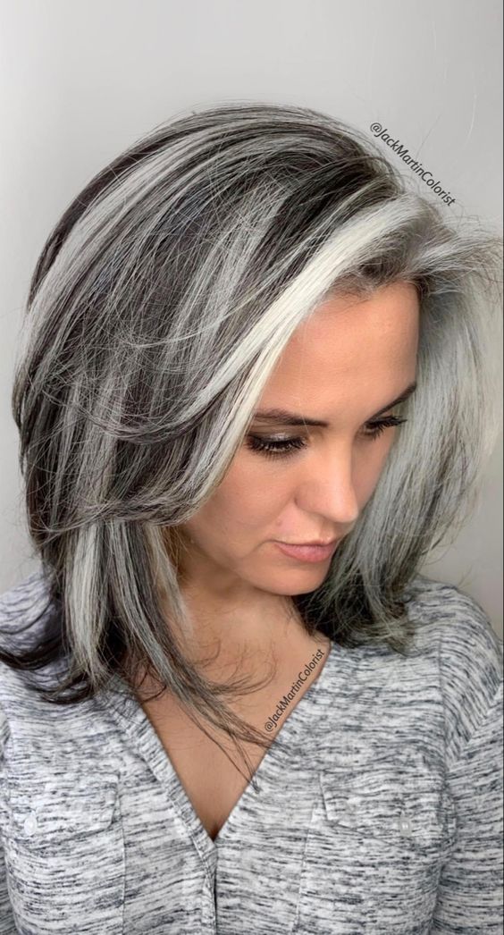 Pessoa com cabelo grisalho elegante e volumoso, vestindo blusa cinza, olhando para baixo.
