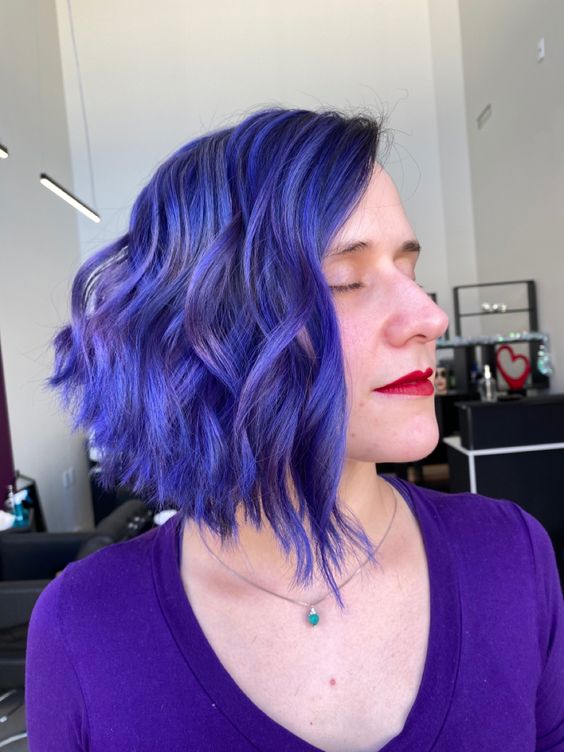 Pessoa com cabelo curto e ondulado em tom azul vibrante, usando blusa roxa, em ambiente interno iluminado.