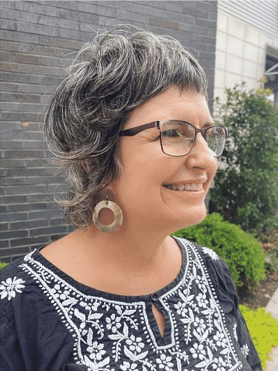Pessoa sorridente com cabelo curto grisalho, usando óculos e brincos grandes, em ambiente externo com plantas ao fundo.