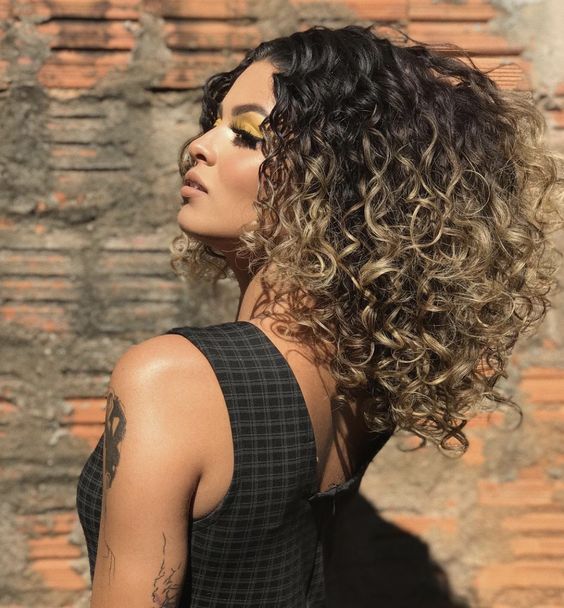 mechas-balayage-cachos-pinterest Pessoa com cabelo cacheado e maquiagem amarela, de perfil, em frente a uma parede de tijolos.