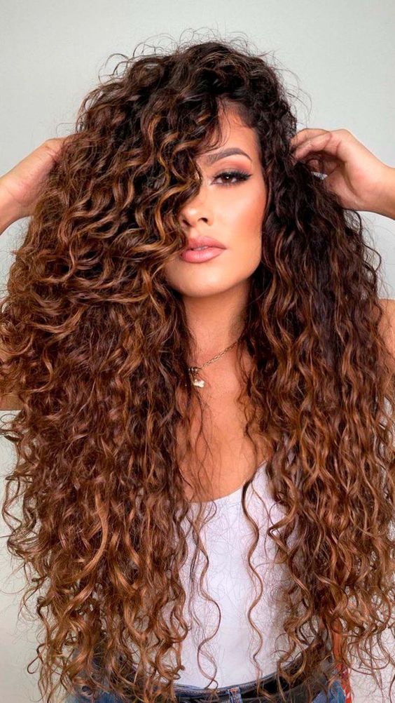 mechas-acobreadas-em-cachos-pinterest Pessoa com cabelos longos e cacheados, vestindo uma blusa branca, em um fundo neutro.