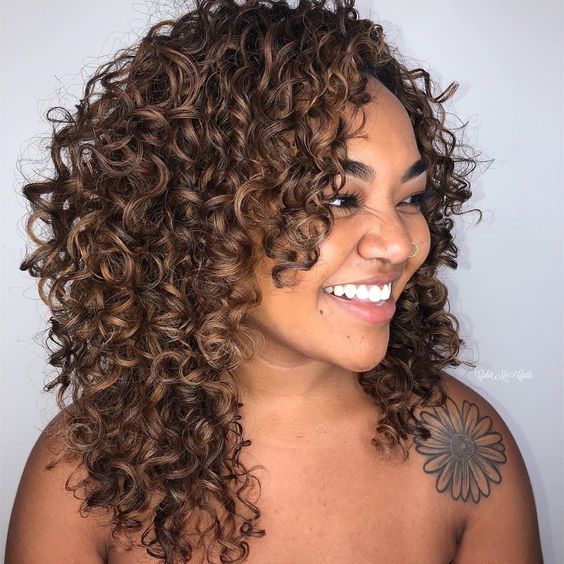 mechas-babylight-em-cachos-pinterest Mulher sorrindo com cabelo cacheado castanho e tatuagem floral no ombro.