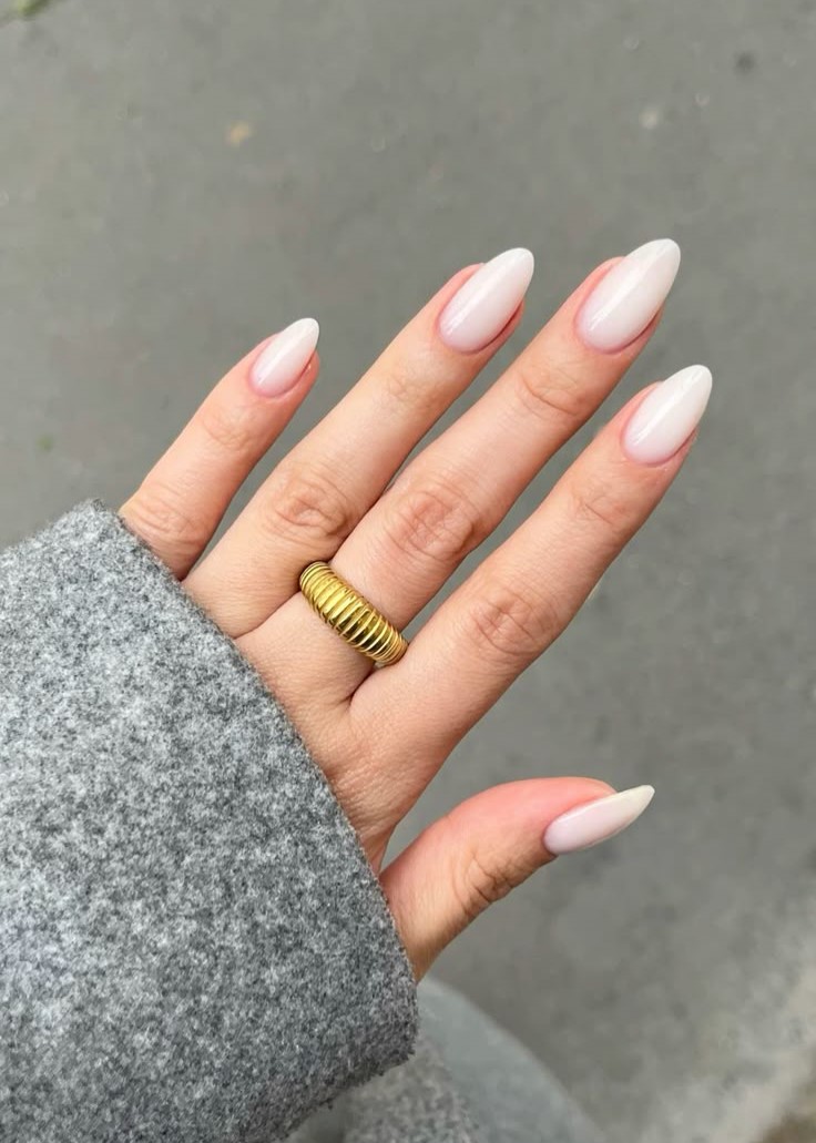 milky-nails-e-tendencia-para-2025-1 Mão com unhas longas e pintadas de nude, usando anel dourado, sobre fundo cinza e vestindo casaco cinza.