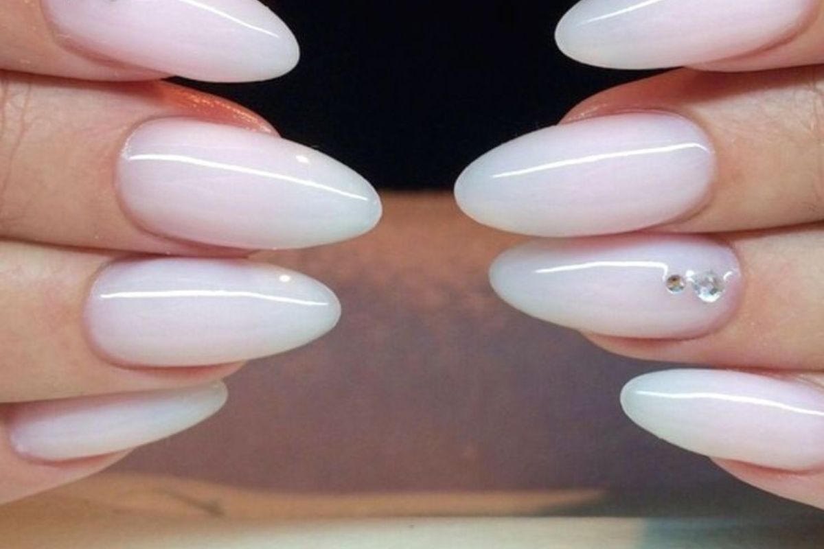 Unhas com esmalte rosa claro, formato amendoado, detalhe de pedrinhas decorativas em uma delas.