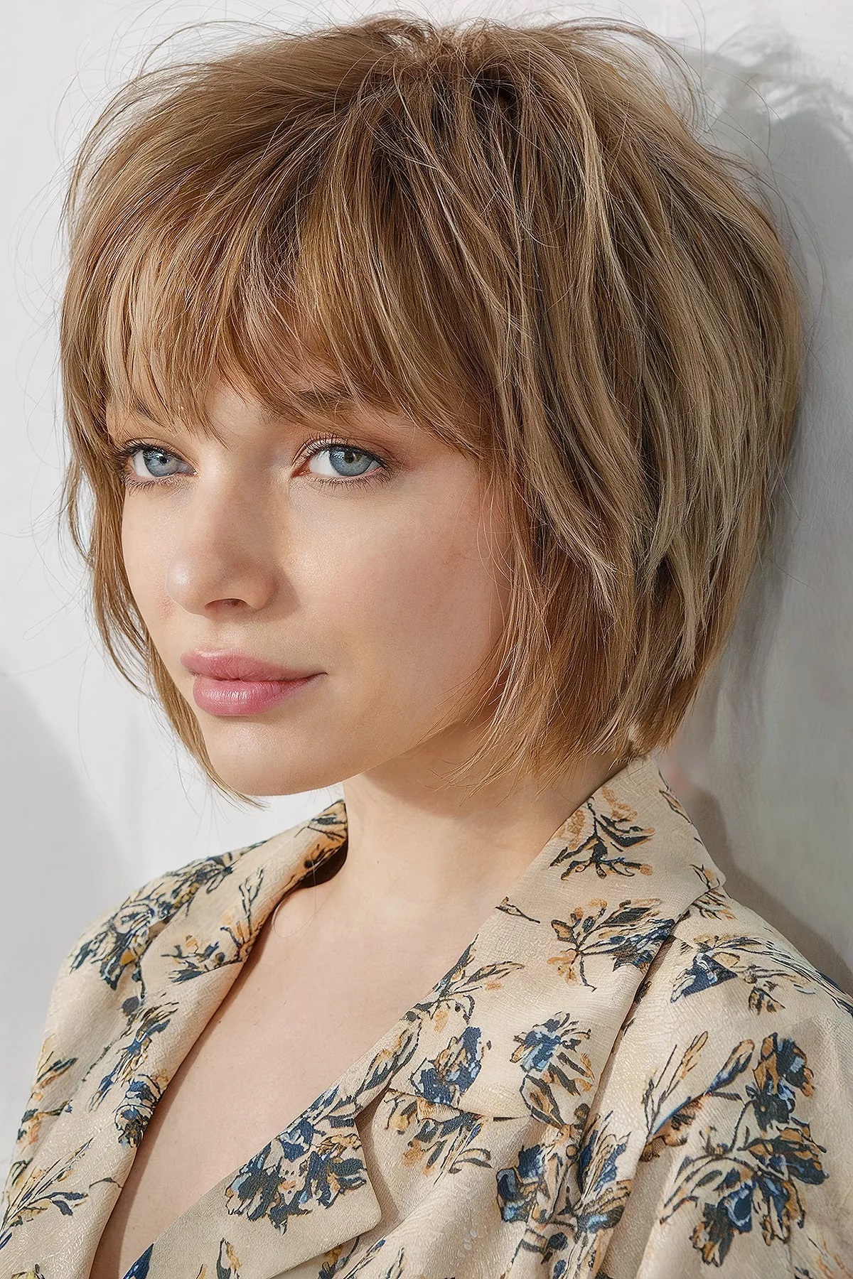 Pessoa com cabelo curto e loiro, vestindo uma blusa estampada com flores em tons de azul e bege, olhando para o lado.