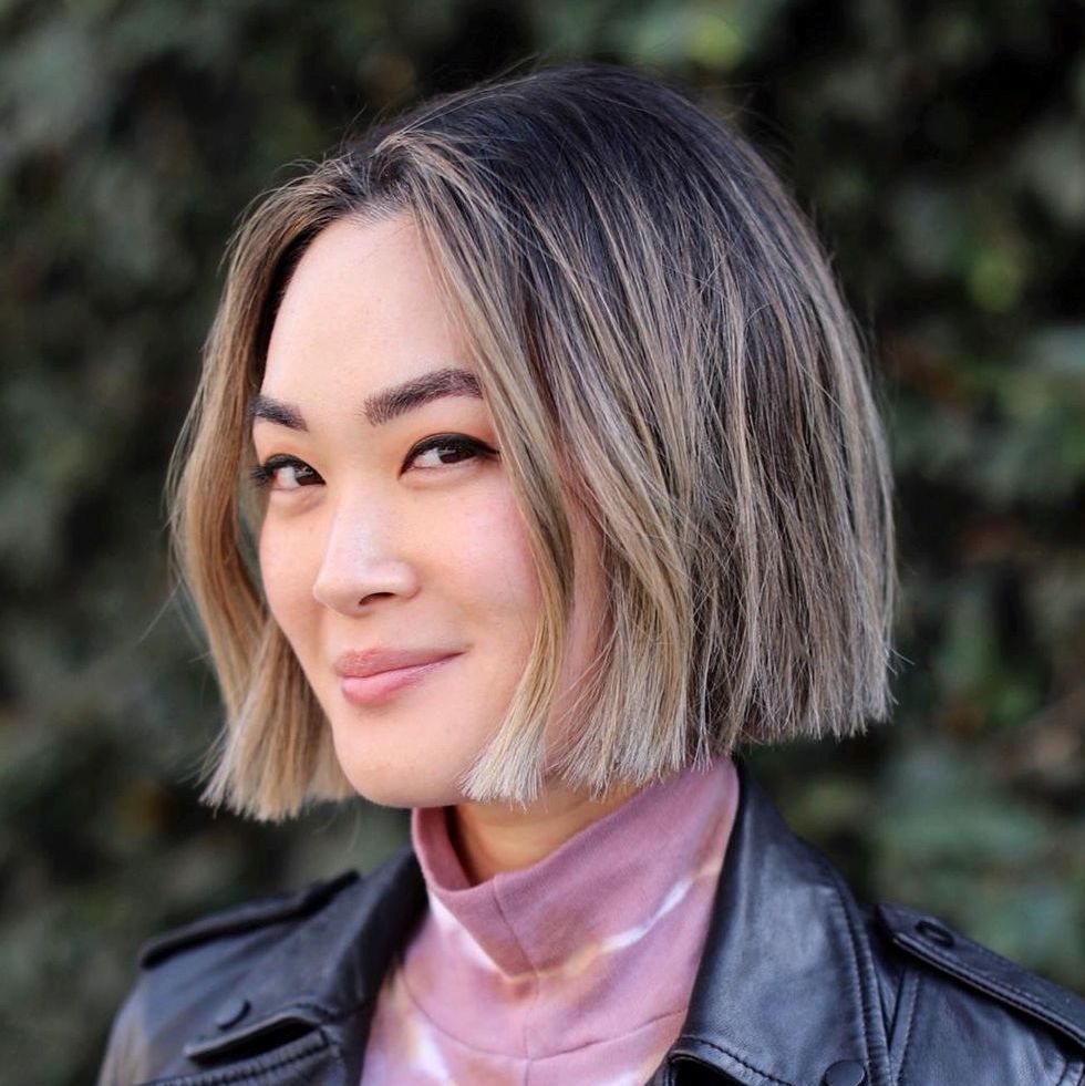 Pessoa sorrindo, com cabelo curto e loiro, usando jaqueta preta e blusa rosa, fundo desfocado de folhagens.