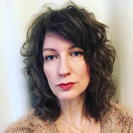 corte-shag-pinterest Mulher de cabelo ondulado castanho, usando suéter bege, em frente a fundo claro.
