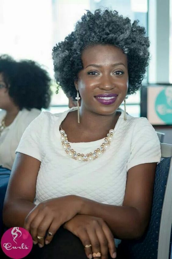 bob-assimetrico-verao-pinterest Pessoa sorridente com cabelo cacheado grisalho, vestindo blusa branca e colar de pérolas, sentada em cadeira.