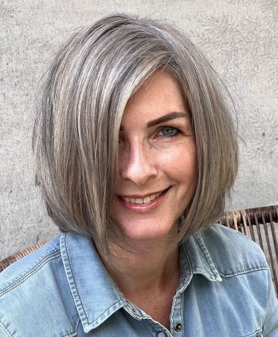 mechas-loiro-acinzentado-pinterest Pessoa sorrindo com cabelo grisalho e curto, vestindo camisa jeans, sentada em uma cadeira de madeira.