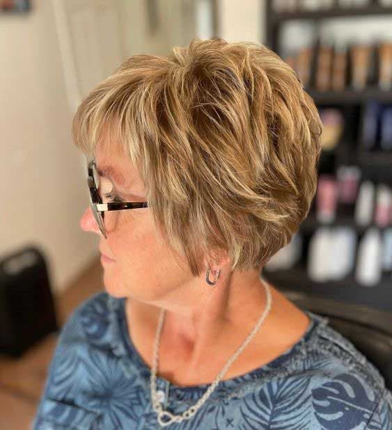 mechas-balayage-para-mulheres-de-50-anos Pessoa de perfil com cabelo curto loiro, usando óculos e blusa estampada, em ambiente interno.