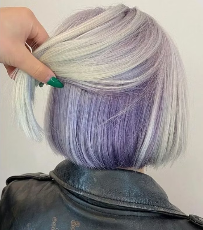 Cabelo curto tingido de roxo e cinza, sendo levantado por uma mão com unhas pintadas de verde.