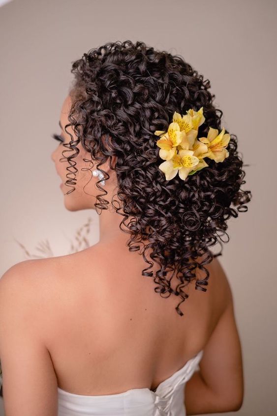 Pessoa com cabelos cacheados presos, adornados com flores amarelas, vestindo traje branco, vista de costas.